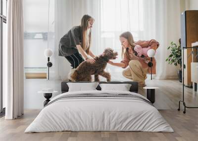 Little girls play with fluffy brown Maltipoo dog in bright, sunlit room. One girl holds stuffed toy. They playing with dog creating joyful and lively scene of indoor playtime and companionship Wall mural