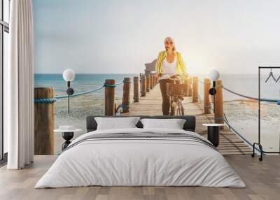 Portrait of a happy smiling woman dressed in light summer clothes and sunglasses riding a bicycle on the wooden sea pier and looking at camera. Careless vacation in tropical countries concept image Wall mural