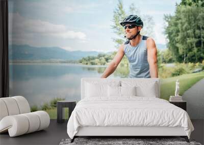 Portrait of a sincerely smiling man dressed in cycling clothes, helmet and sunglasses riding a bicycle on the asphalt out-of-town bicycle path along a mount lake. Active sporty people concept image. Wall mural