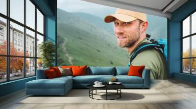 Portrait of backpacker man in baseball cap walking by the foggy cloudy weather mountain range path with backpack. Active sports backpacking healthy lifestyle concept. Wall mural