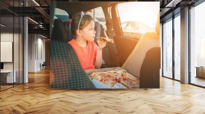 Portrait of little 5 YO girl eating just cooked Italian pizza sitting in child car seat on car back seat and looking out window . Happy childhood, fast food eating, or auto journey lunch break image Wall mural