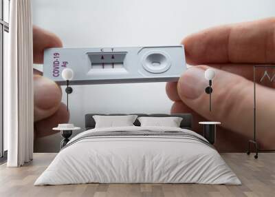 Rapid antigen detection test (RADT) with two red stripes showing a POSITIVE result of a human sample testing. Mans fingers holding the white plastic device with a COVID-19 Ag inscription. Wall mural