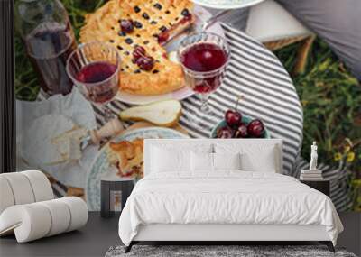 Served table top view with fruit pie and vintage tableware, red wine in glasses, man eating a tasty dessert,  sitting in rattan chairs on the high grass meadow next the table and enjoying a tart. Wall mural