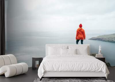 Trail runner female dressed orange sporty hoodie and red cap resting on the cliff and enjoying Atlantic ocean view on Ponta de Sao Lourenço peninsula -the easternmost point of Madeira island, Portugal Wall mural