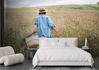 Young Woman dressed jeans jacket and light summer dress walking by the high green grass meadow with basket and wildflowers bouquet. Human and nature concept back shot photo. Wall mural