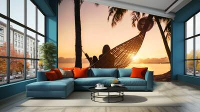 Young woman relaxing in hammock hinged between palm trees on the sand beach at orange sunrise morning time Wall mural