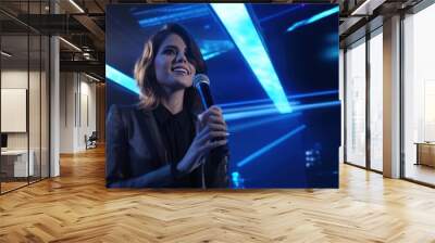 A female emcee in a suit and glasses stands on stage looking at the camera and announcing the opening of a musical performance. In a bright room with LED screen and night light Wall mural