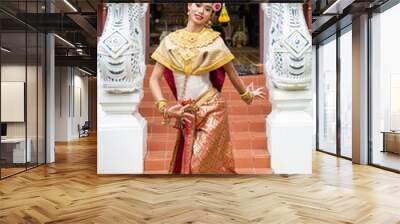 A traditional Thai dancer in a gold and red ornate costume poses with hands together in front of golden temple. Wall mural