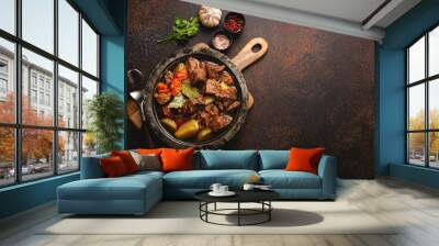 Delicious beef meat stew dish with potatoes, carrot and gravy in rustic metal bowl with spoon, bunch of fresh parsley, garlic cloves, spices on brown concrete background top view space for text Wall mural