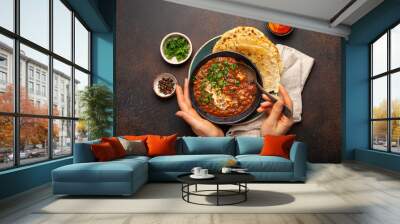 Female hands holding a bowl and eating traditional Indian Punjabi dish Dal makhani with lentils and beans served with naan flat bread, fresh cilantro on brown concrete rustic table top view. Wall mural