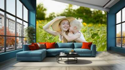 Happy laughing mother giving toddler son piggyback ride on background of green blooming sunflowers field Wall mural