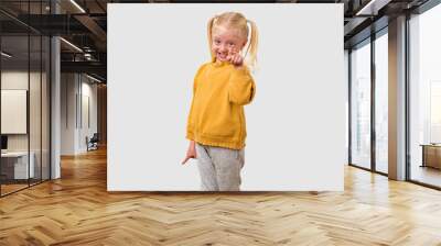 Portrait of a cute little girl with blond hair, dressed in a yellow sweater and gray pants, showing thumbs up gesture, isolated over white background. Wall mural