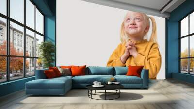 Portrait of a little girl with blond hair in a yellow sweater standing with her arms crossed in prayer and looking up on a white background. Wall mural