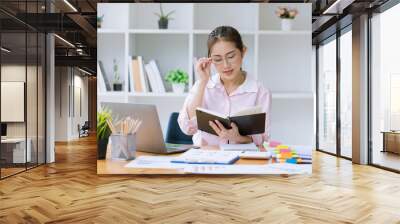 Young asian woman wear glasses reading a book with laptop sitting at desk in the study room. Wall mural