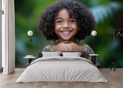 A happy young child with curly hair beams while holding a coconut, surrounded by vibrant tropical foliage on a sunny day Wall mural