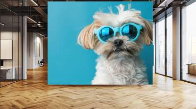 A small dog with fluffy fur and stylish blue sunglasses sits confidently in front of a vibrant blue background, radiating charm Wall mural