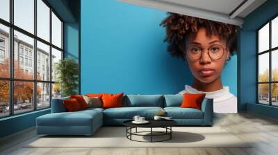 A young woman stands confidently with her arms relaxed, showcasing her curly hair and round glasses against a vivid blue backdrop Wall mural