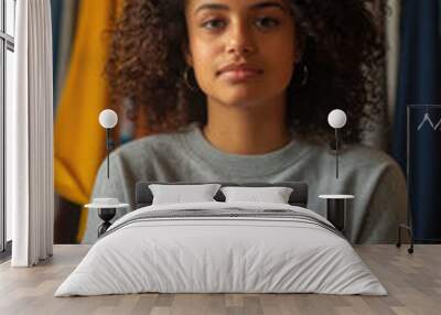 A young woman with curly hair sits calmly amidst a vibrant array of clothing, showcasing her personal style and peaceful demeanor Wall mural