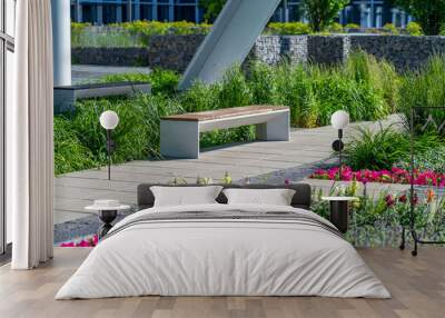 Empty concrete bench with wooden slats for sitting on tile among decorative grass and flowers in recreation area near modern office building. Garden landscape with chair in city park Wall mural