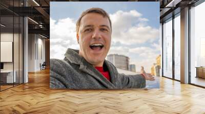 Portrait of millennial man recording video, take selfie photo, looking at camera, smiling wide, showing on city background Wall mural