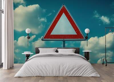 A red triangular warning sign against a bright blue sky filled with clouds, indicating caution for drivers. Wall mural