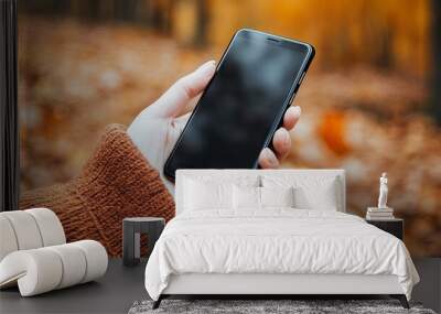 Close-up of a woman's hand holding a smartphone with an autumn forest in the background. Wall mural