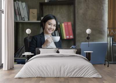 A pretty Asian businesswoman holding a coffee cup while sitting at her office desk. Wall mural