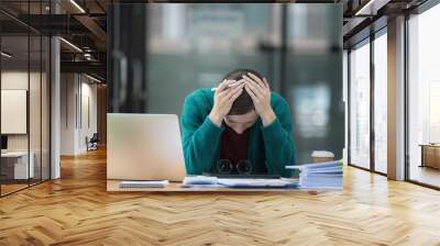 A tired businesswoman under stress works at a laptop while sitting at a table at home and holds her hand on her temples, migraine attacks. Wall mural