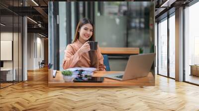 Beautiful young Asian businesswoman drinking a coffee while working on a laptop at the office. Looking at the camera.
 Wall mural