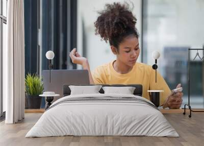 Portrait of a beautiful confident businesswoman using a laptop computer holding a mobile phone sitting in a modern office. Smiling African American freelancer working online from home. Wall mural