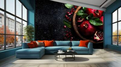   A wooden bowl brimming with luscious pomegranates atop a black table, surrounded by vibrant foliage Wall mural