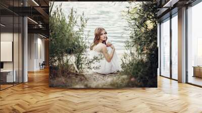 beautiful boho girl sitting on sandy beach at sunset light near lake. attractive young woman in white bohemian dress with windy hair relaxing  near water among green leaves. summer vacation Wall mural