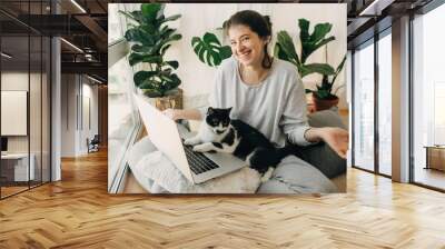 Cute cat helping owner during quarantine, loyal companion. Casual girl working on laptop with her cat, sitting together in modern room with pillows and plants. Home office.  Stay home stay safe Wall mural