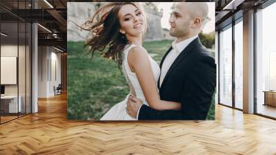 Gorgeous bride and groom having fun and dancing in evening sunlight near old castle in park. Stylish wedding couple gently embracing  and smiling. Romantic moment Wall mural