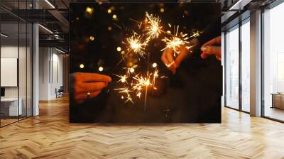 Happy New Year! Friends celebrating with burning sparklers in hands against christmas tree lights in dark room. Hands holding fireworks on background of stylish decorated illuminated tree. Moody Wall mural