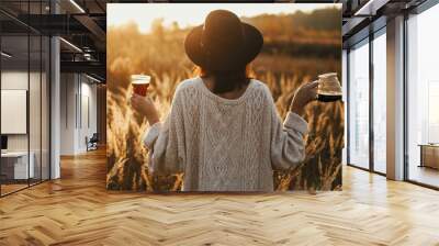 Hipster woman in hat holding hot coffee and glass cup on background in sunny warm light in rural herbs. Alternative coffee brewing. Atmospheric rustic tranquil moment. Back view Wall mural