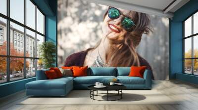 Portrait of happy boho girl in cool outfit and sunglasses smiling in sunny street. Stylish hipster carefree girl posing on background of wooden wall. Space for text. Summer vacation and travel Wall mural