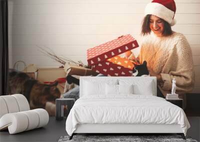 Stylish happy girl in santa hat and her cats opening christmas gift box with magic light in decorated christmas room. Young hipster woman opening magical present. Merry Christmas Wall mural