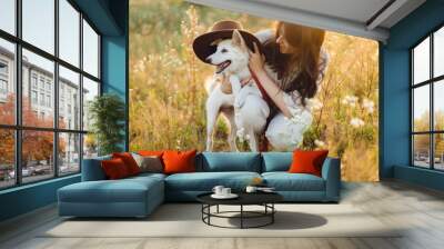 Stylish happy woman playing with cute dog with hat among wildflowers in sunset light. Summer travel with pet. Young carefree female having fun with white danish spitz in summer meadow Wall mural