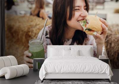 Stylish hipster girl in sunglasses eating delicious vegan burger and holding smoothie in glass jar in hands at street food festival. Happy boho woman biting burger with drink in summer street Wall mural