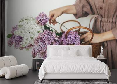 Stylish woman with beautiful lilac flowers in wicker basket in rustic room. Female in linen dress arranging lilac flowers in rural room, cropped view. Authentic spring moments Wall mural