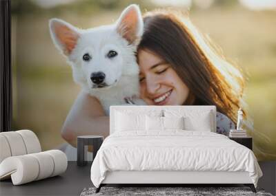 Woman hugging cute white puppy in warm sunset light in summer meadow, sweet authentic moment. Happy girl holding adorable fluffy puppy close up. Adoption concept Wall mural