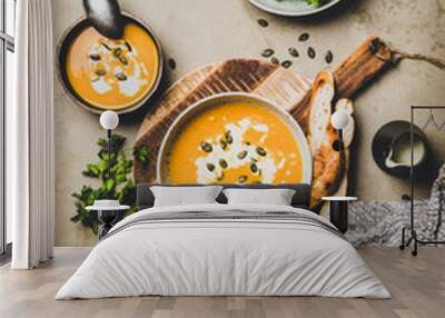 Autumn, winter warming seasonal dinner. Flat-lay of pumpkin soup with seeds, parsley, cream and bread in bowls over grey concrete background, top view. Vegan, vegetarian, healthy, comfort food concept Wall mural