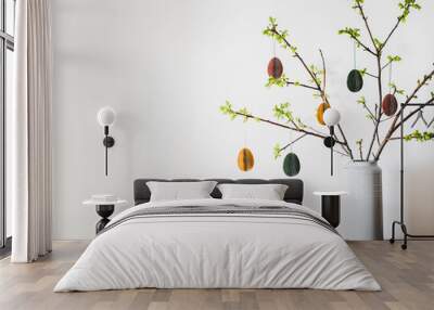 Easter holiday home decoration. Tree branches in vase with fresh spring leaves decorated with festive colorful Easter eggs, white wall background, copy space. Easter holiday preparation, greeting card Wall mural