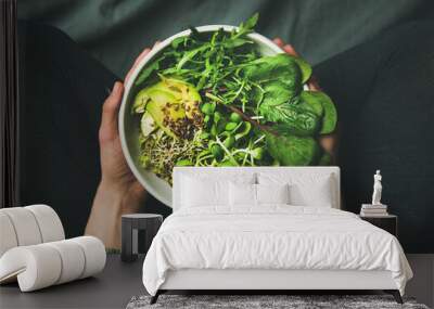 Green vegan breakfast meal in bowl with spinach, arugula, avocado, seeds and sprouts. Girl in leggins holding plate with hands visible, top view. Clean eating, dieting, vegan food concept Wall mural