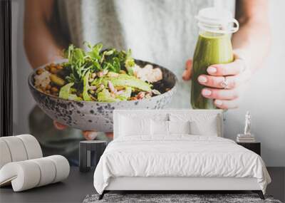 Healthy dinner or lunch. Woman in t-shirt and jeans standing and holding vegan superbowl or Buddha bowl with hummus, vegetable, salad, beans, couscous and avocado and smoothie in hands, square crop Wall mural