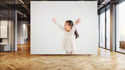 A jubilant kid girl raises her fists in celebration of her success, saying yes with excitement. Asian portrait of a happy young kindergarten child in a studio shot on a white background, winning Wall mural