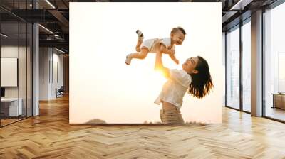 A woman holds her newborn baby up high, as they enjoy a moment of nature in the park at sunset. The little one looks up at the sky with wonder, while the proud mom captures the precious life moment Wall mural