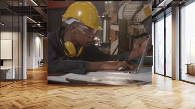 American industrial black young worker man with yellow helmet and ear protection typing keyboard of laptop computer in front machine, Confident engineer at work in the industry factory Wall mural