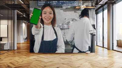 Asian happy beautiful young woman standing behind the bar counter in cafe smiling showing smartphone blank green screen looking camera, small business owner coffee shop Wall mural